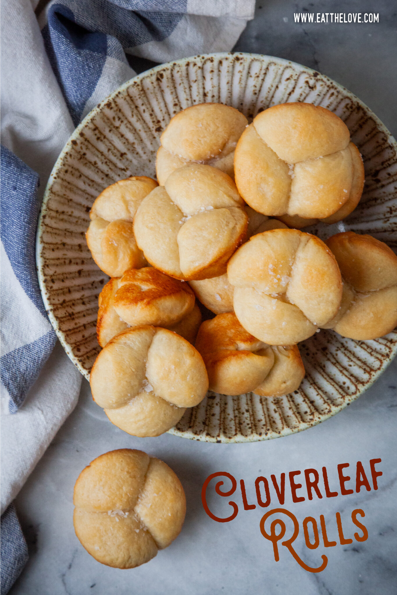 A bowl of cloverleaf shaped bread rolls, with one roll on the counter. A blue and white cloth napkin is next to the bowl. The type on the image says Cloverleaf Rolls.