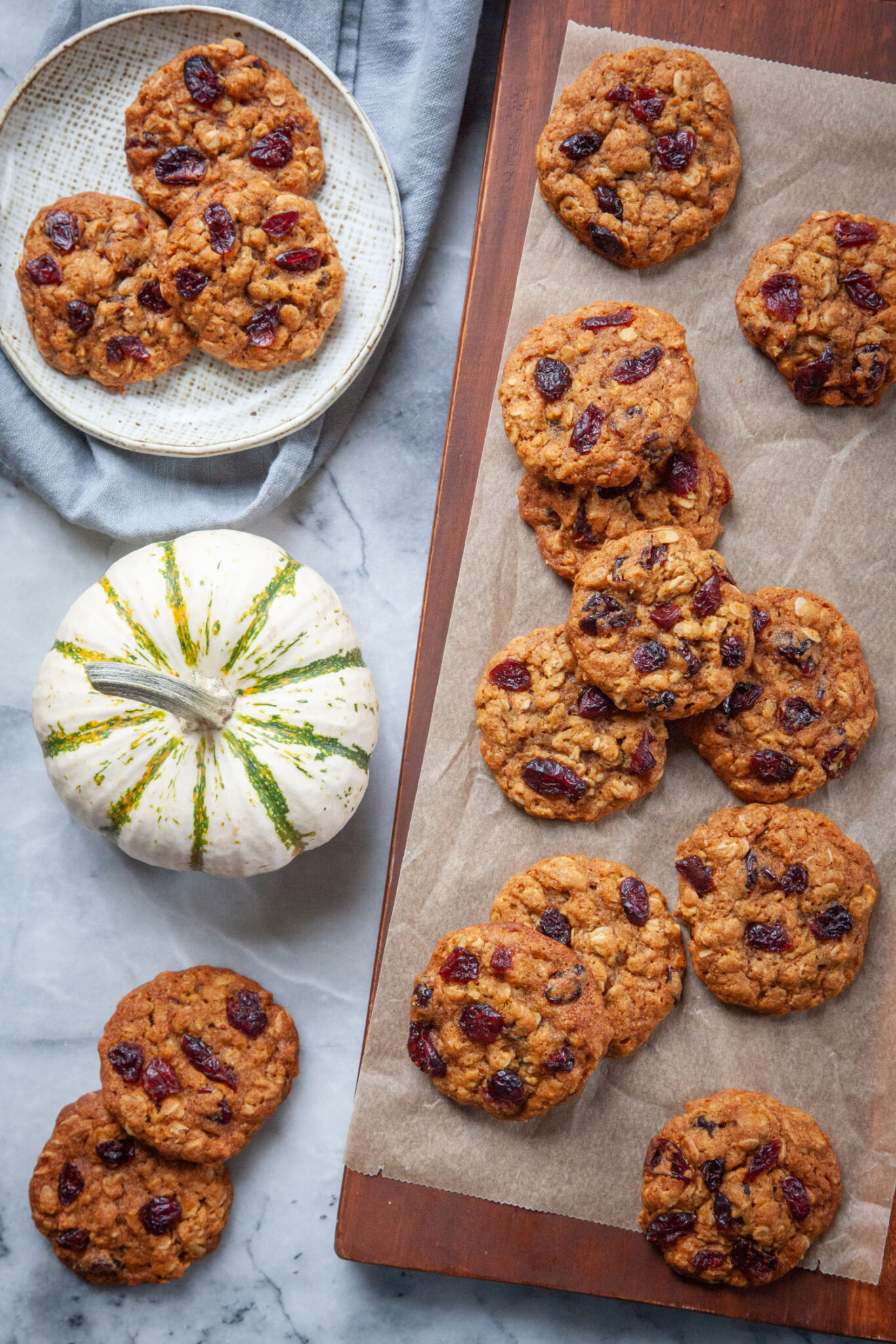 Pumpkin oatmeal cranberry cookies on a wooden cutting board, with more cookies on a plate and on the table. A small decorative pumpkin is next to the cutting board.