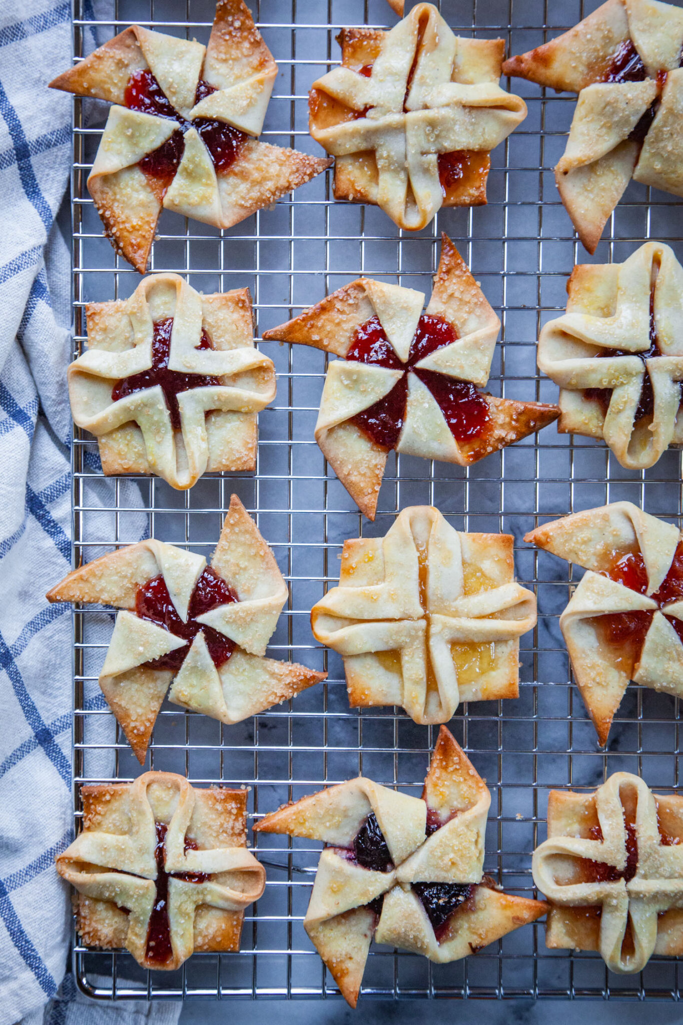 Finnish Christmas Cookies, called Joulutorttu, on a wire rack with a cloth napkin next to them. The cookies are jam filled pastries that are formed to look like a square flower and in a pinwheel shape.