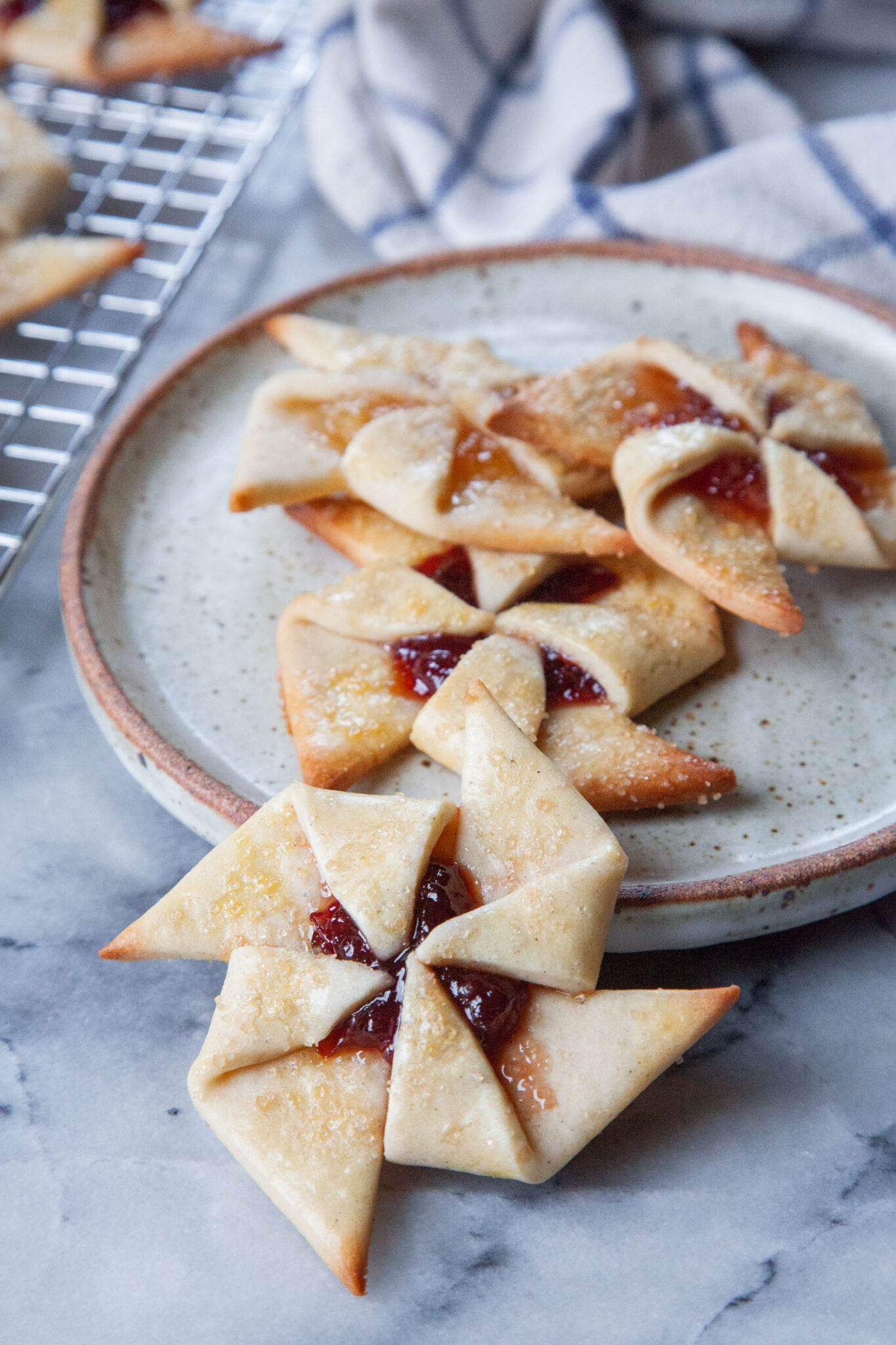 A plate full of joulutorttu cookies shaped like pinwheels on a plate. There is a wire rack with more cookies behind the plate.