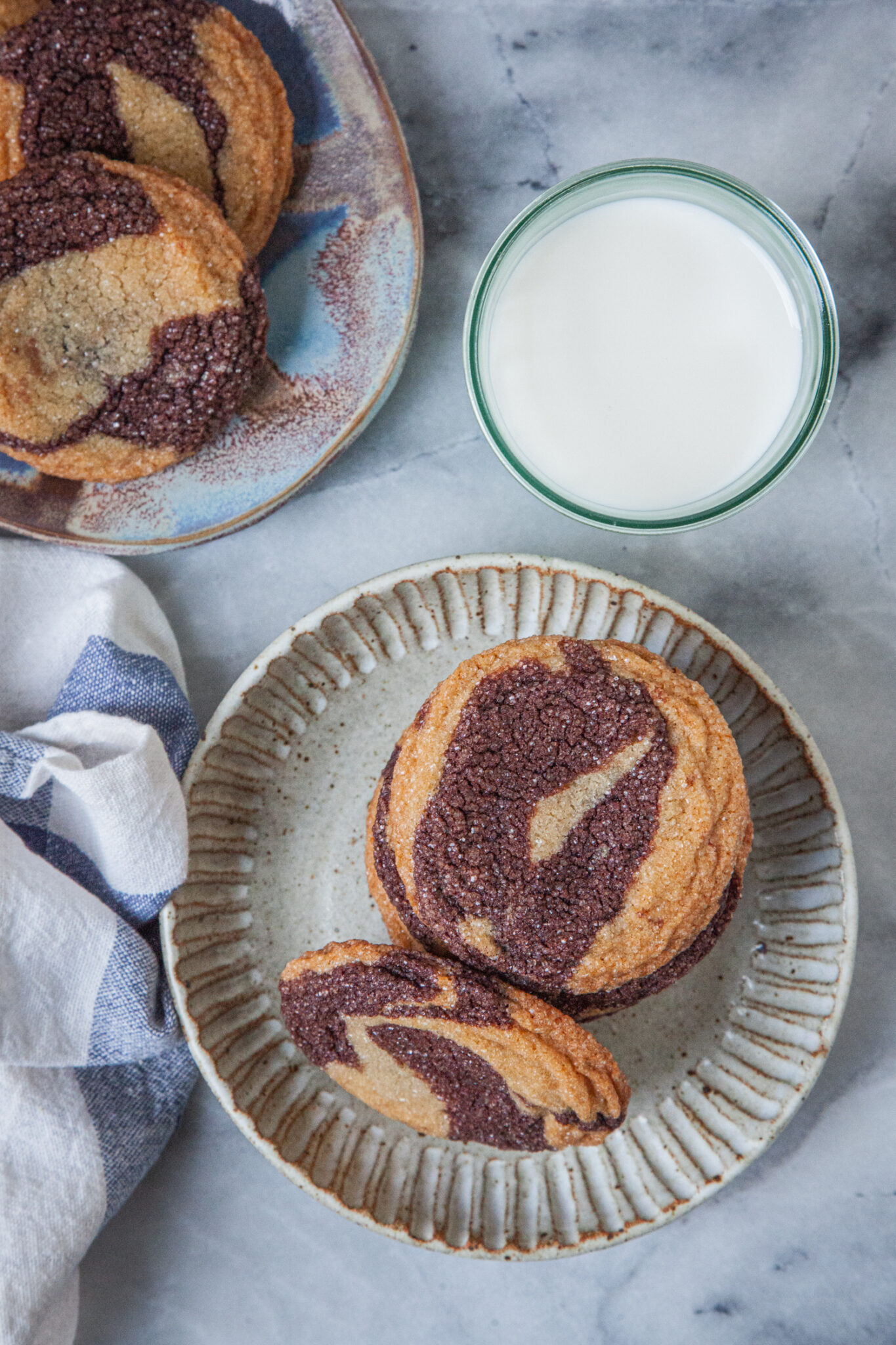 A stack of marbled sugar cookies in a plate, with a glass of milk next to it, and more cookies on a plate next the glass.