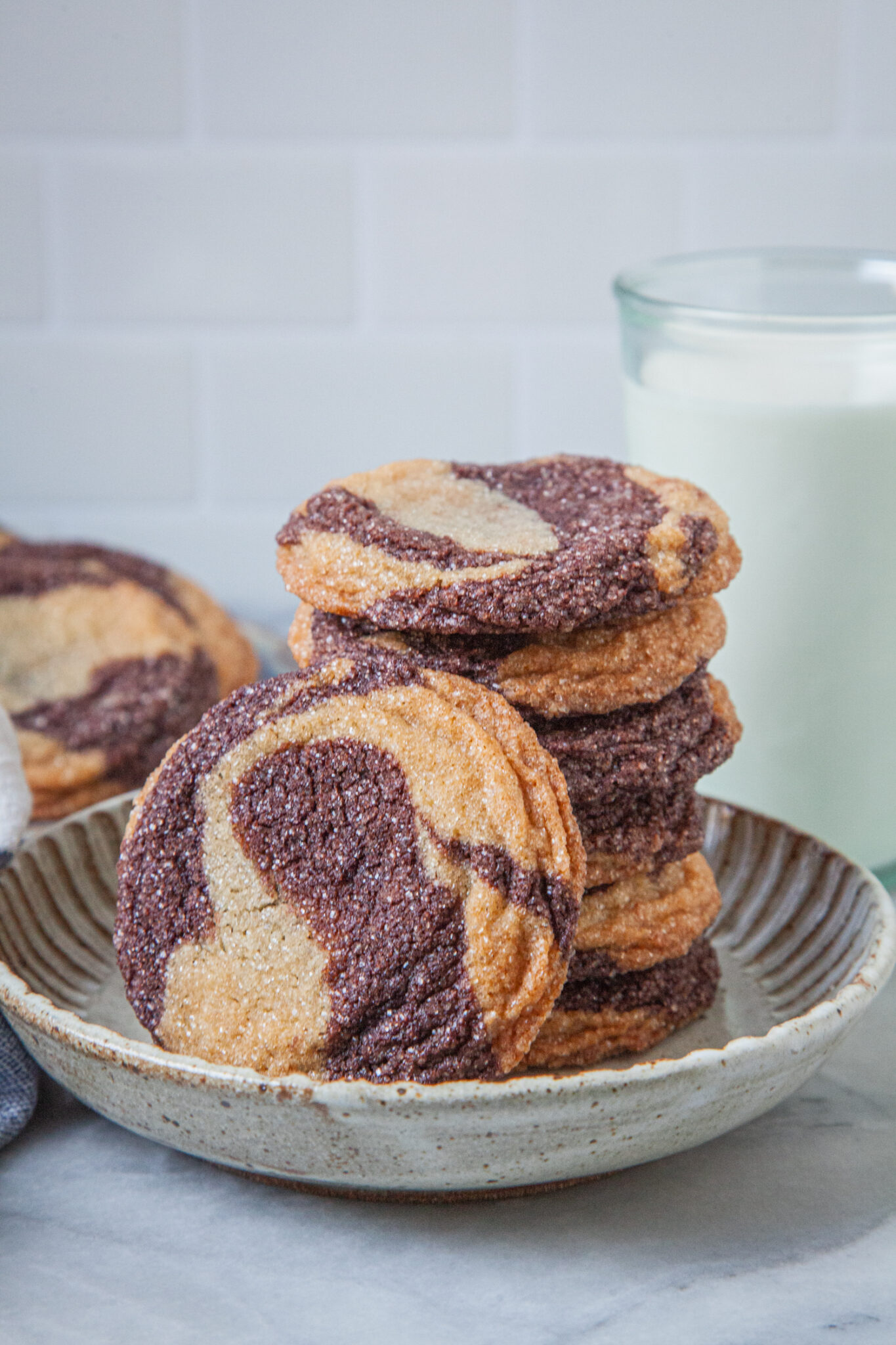 A stack of marbled sugar cookies on a plate, with a glass of milk behind the cookie and more cookies behind the plate. 