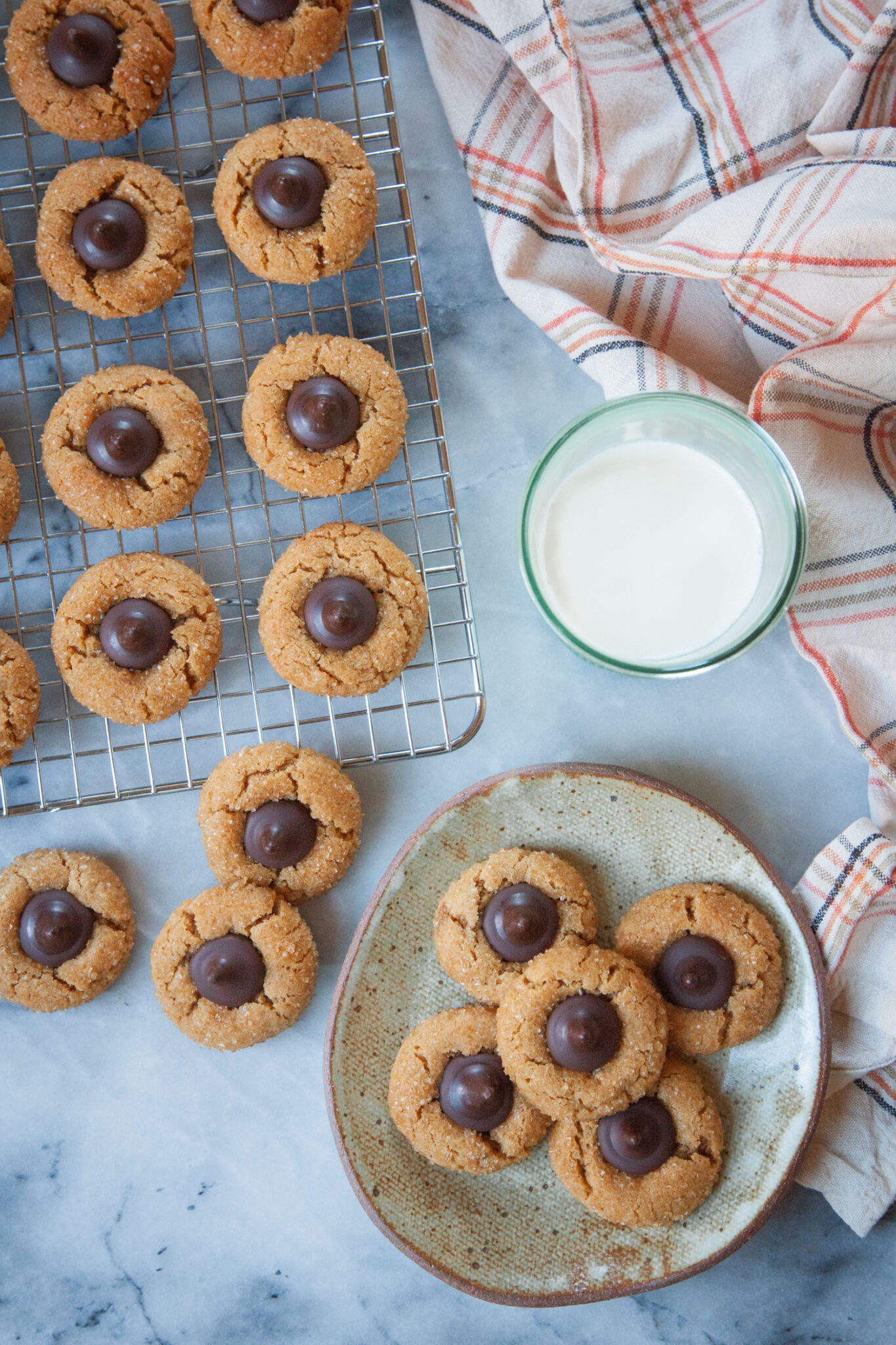 A small plate of peanut butter blossom cookies with a wire cooling rack filled with more cookies. There is a glass of milk and a cloth napkin next to the wire rack and plate. 