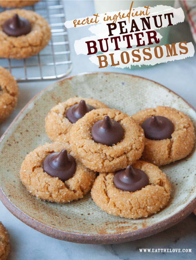 A plate with peanut butter blossom cookies on it. There is a glass of milk along with a wire cooling rack with more cookies on it behind the plate.
