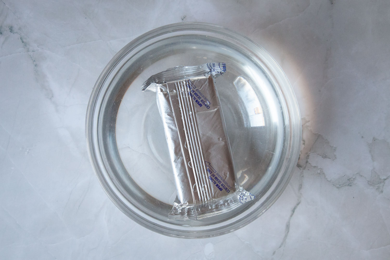 A brick of cream cheese still in its foil wrapper, submerged in hot water in a glass bowl.