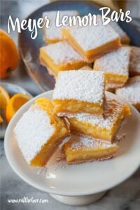 A stack of Meyer lemon bars on a small elevated plate, with more Meyer lemon bars behind it. There is a small plate of Meyer lemon wedges and a whole Meyer lemon behind it.