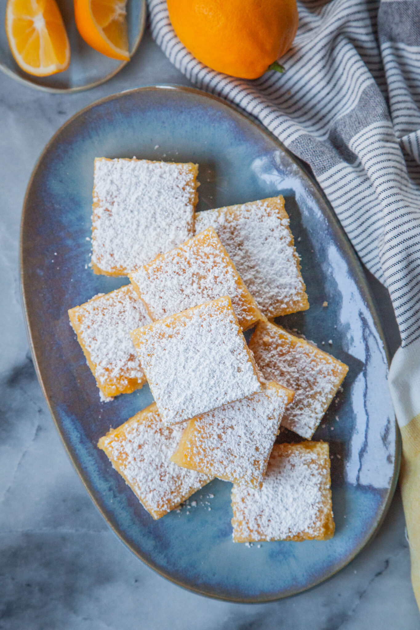 Meyer lemon bars on a blue oval plate, with wedges of Meyer lemon on a small plate next to it, along with a whole Meyer lemon. 