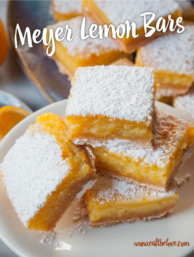 A stack of Meyer lemon bars on a small elevated plate, with more Meyer lemon bars behind it. There is a small plate of Meyer lemon wedges and a whole Meyer lemon behind it.