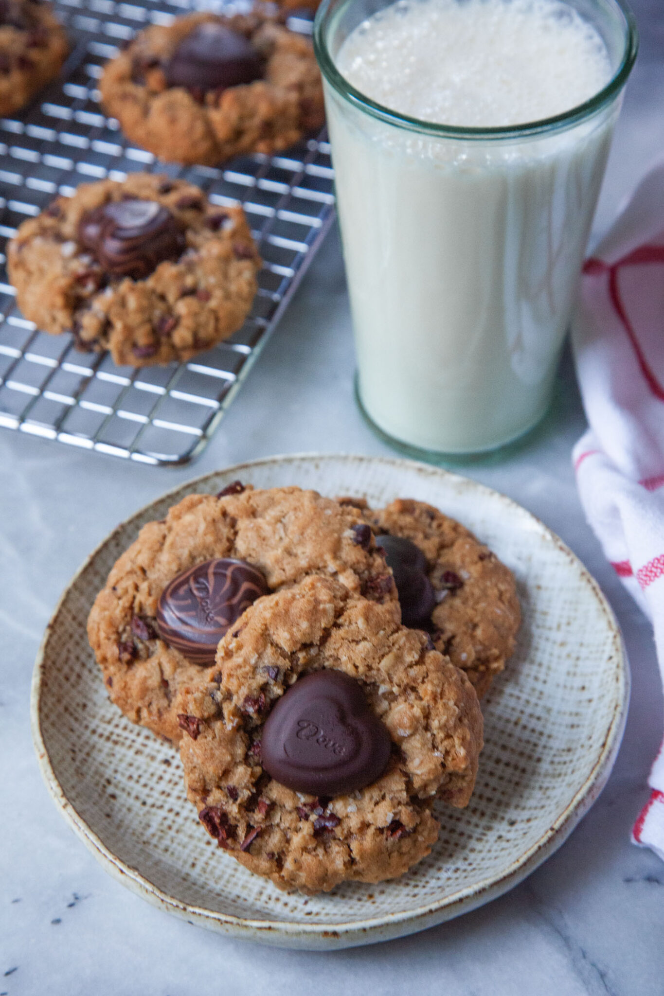 Three oatmeal dried strawberry cookies with a chocolate heart on a plate. There is a glass of milk and more cookies on a wire cooling rack behind the plate.