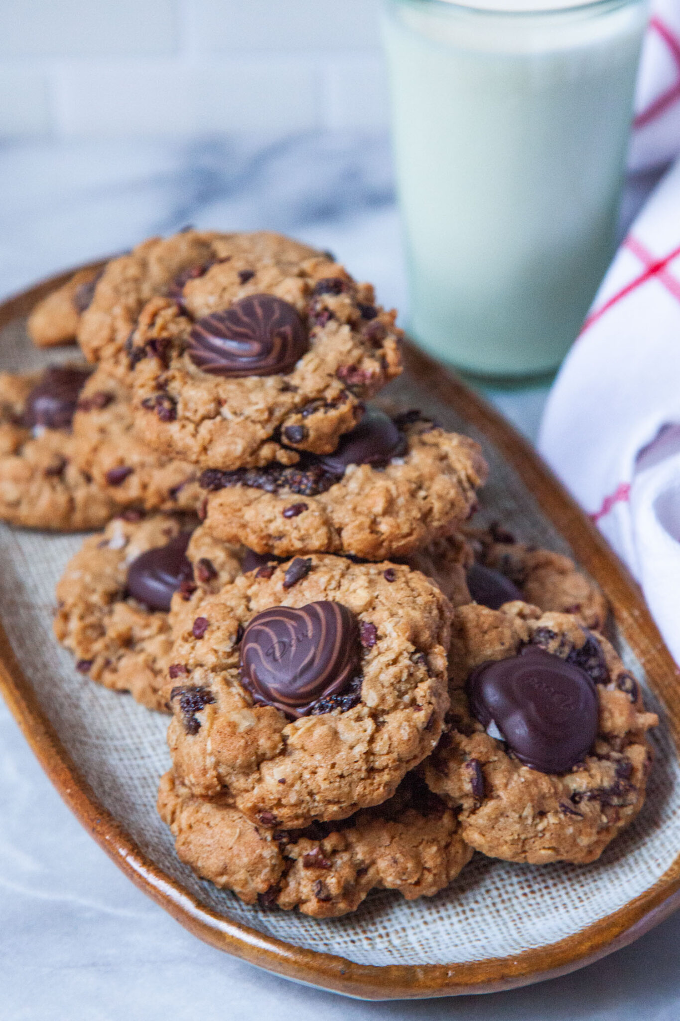 A long oval plate filled with oatmeal dried strawberry cookies with a chocolate heart in the center of each cookie. There is a glass of milk and a cloth napkin behind the plate.