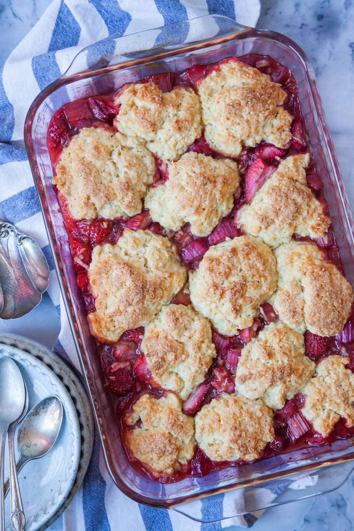 Strawberry Rhubarb Cobbler with fluffy biscuits in a glass casserole dish sitting on a blue striped cloth napkin.