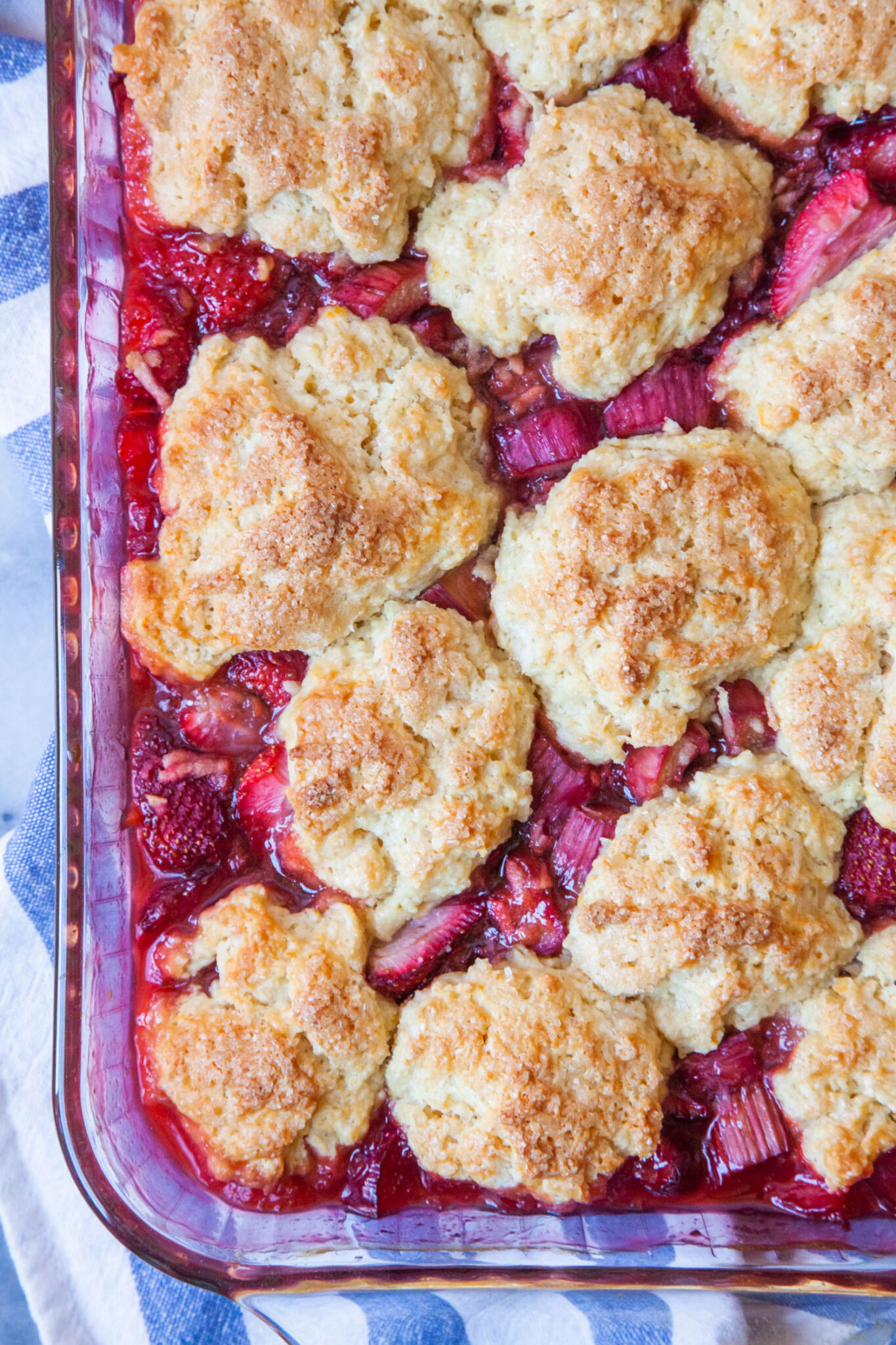 Strawberry Rhubarb Cobbler with fluffy biscuits in a glass casserole dish sitting on a blue striped cloth napkin.