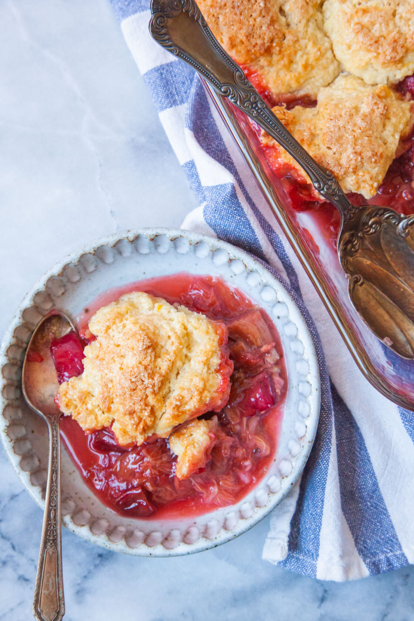 A small bowl with a serving of strawberry rhubarb cobbler in it. The remaining cobbler is behind the bowl.