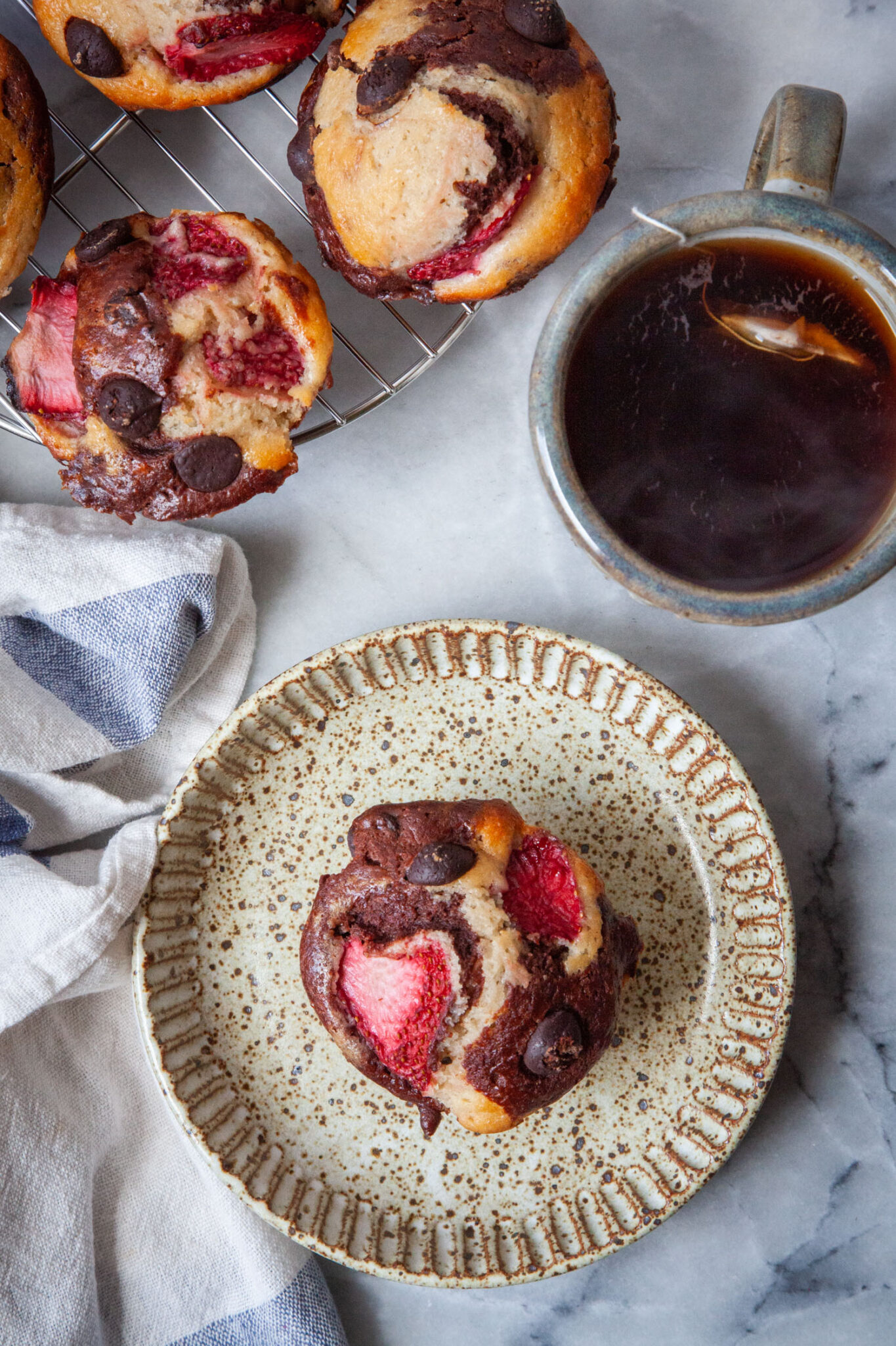 A neapolitan muffin, vanilla, chocolate and strawberry muffin, sitting on a ceramic plate, with more muffins next to it along with a mug of tea. 