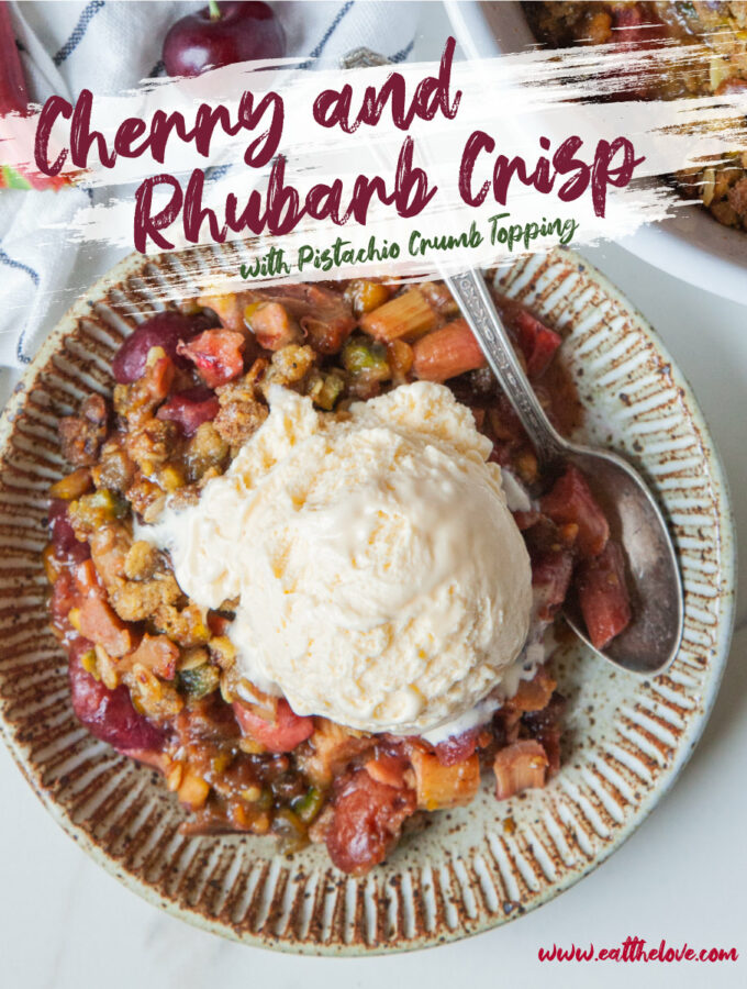 A serving of Cherry Rhubarb Crisp in a plate with a scoop of vanilla ice cream on top. Next to the dish is a casserole dish with the remaining crisp along with some fresh rhubarb and cherries.