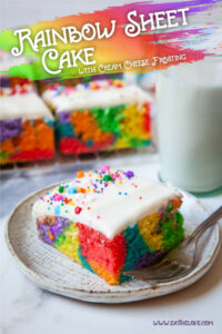 A slice of rainbow sheet cake with cream cheese frosting on a plate. Behind the plate is a glass of milk and more pieces of cake.