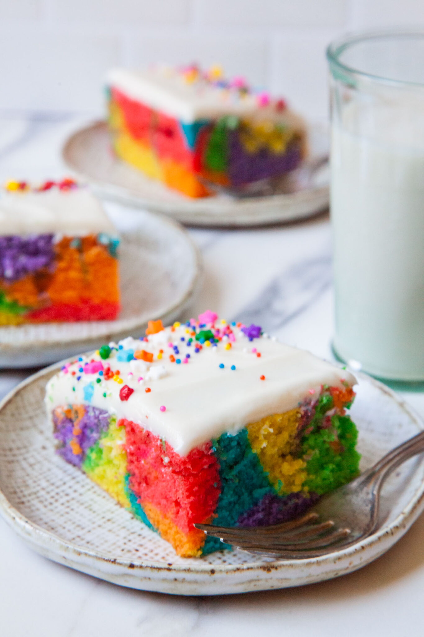 A slice of rainbow sheet cake with a glass of milk behind it, and more pieces of sheet cakes on plates.