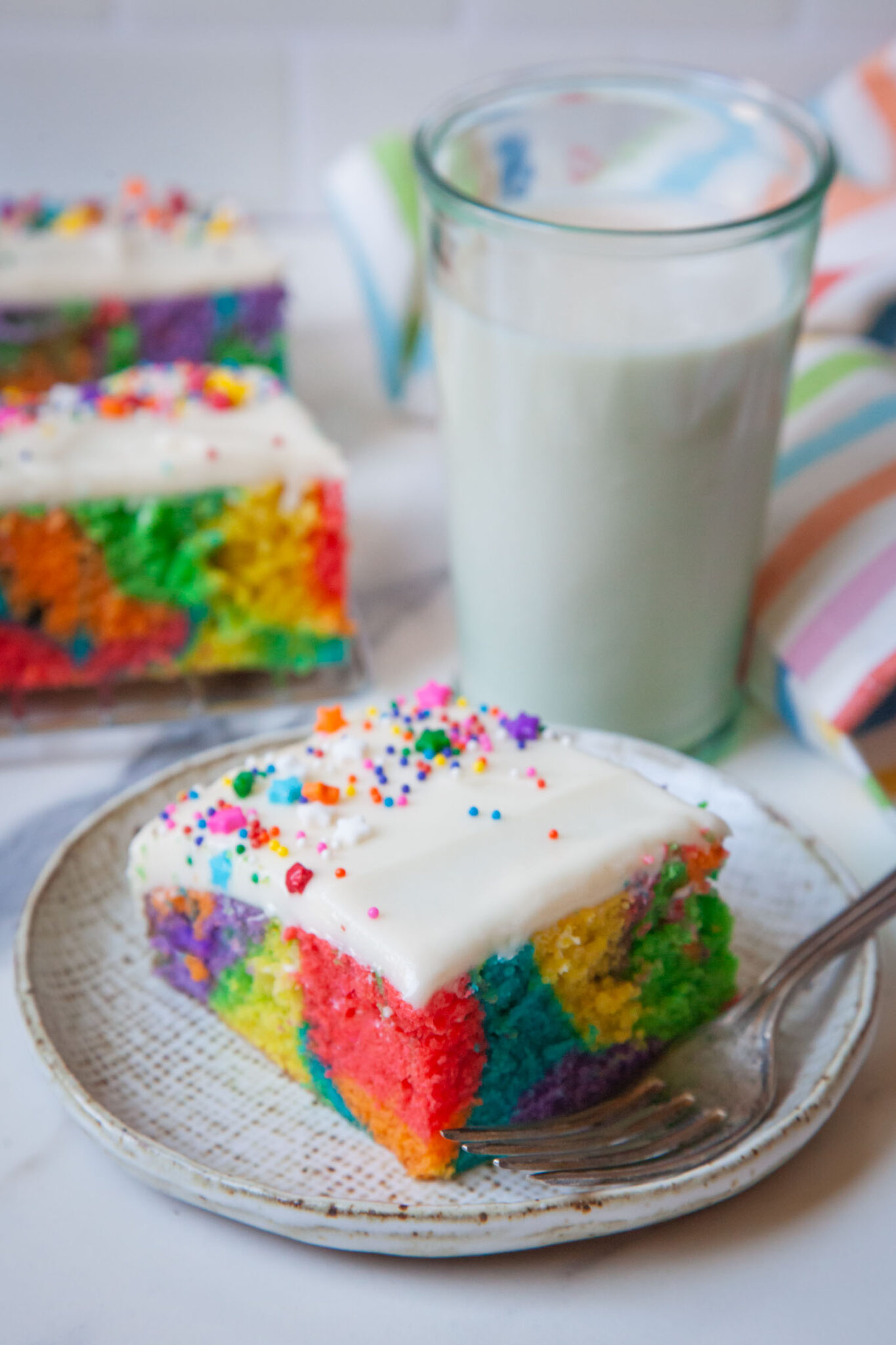 A slice of rainbow sheet cake with a glass of milk behind it, and more pieces of sheet cakes on plates.