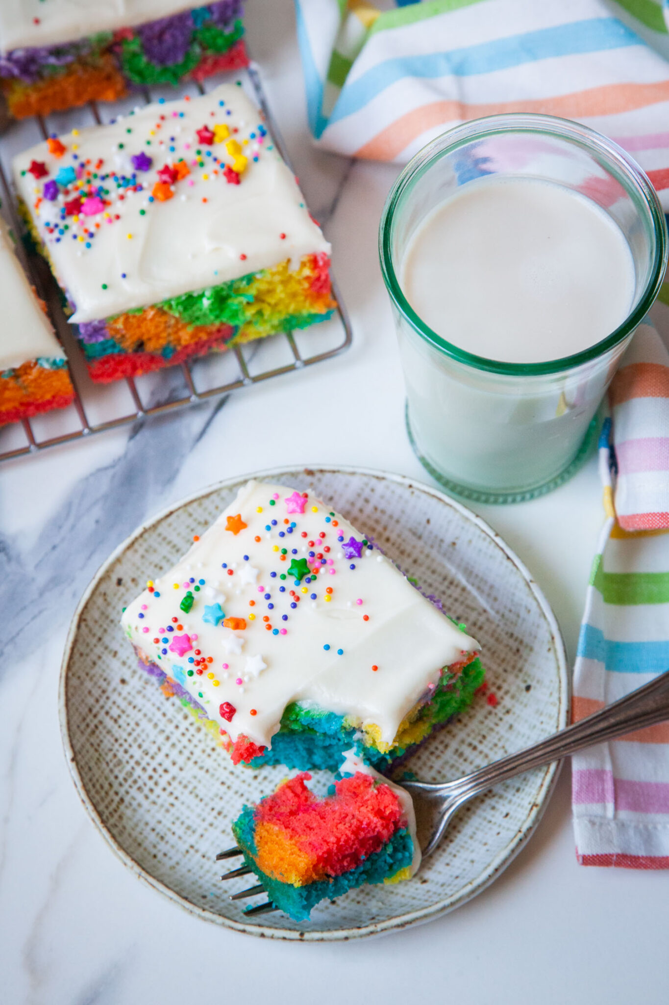 Rainbow sheet cake with a fork that has taken a piece out of the cake. Next to the plate is a glass of milk and more sheet cake pieces on a wire cooling rack.