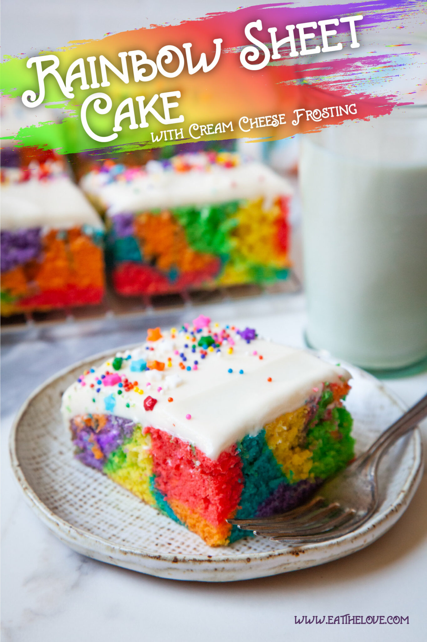 A slice of rainbow sheet cake with cream cheese frosting on a plate. Behind the plate is a glass of milk and more pieces of cake.