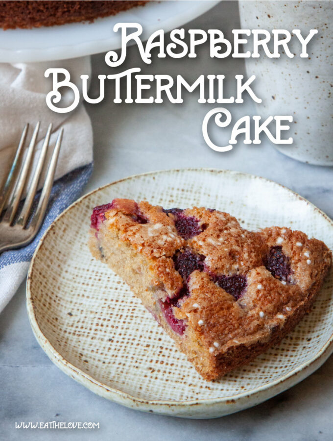 A slice of raspberry buttermilk cake on a plate, with a mug of tea behind it. Behind the tea is the remaining raspberry cake on a cake stand.