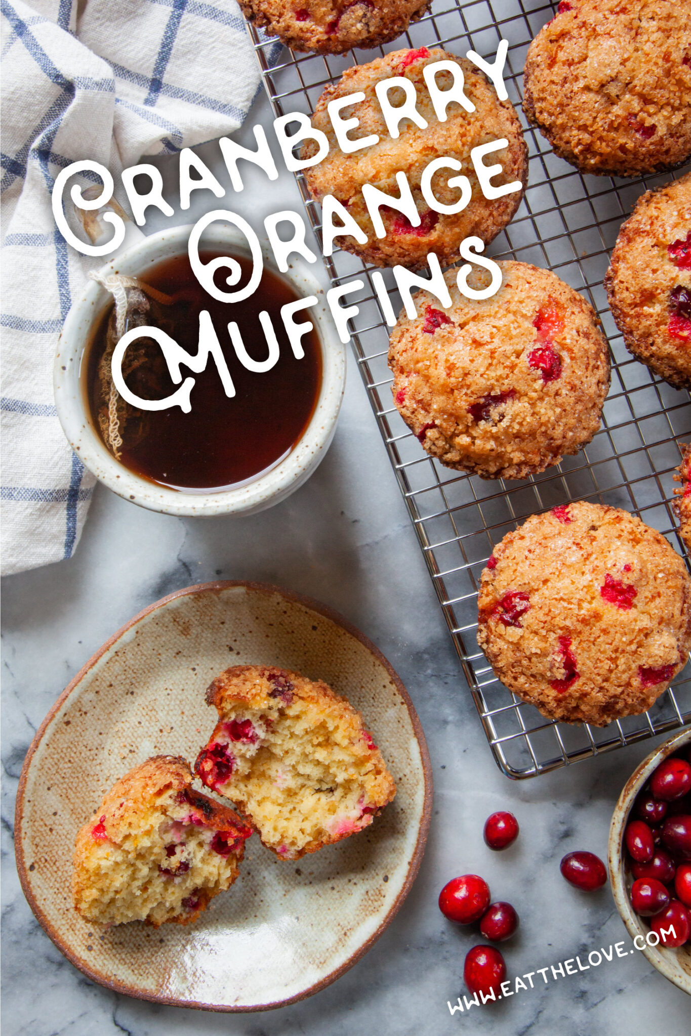 A cranberry orange muffin broken open on a plate, with a mug of tea next to it and more muffins on a wire cooling rack. The text on the image says "Cranberry Orange muffins".