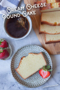 A slice of cream cheese pound cake on a plate with a strawberry garnish. There is a mug of tea next to the plate, along with a wooden serving board with the rest of the cream cheese pound cake loaf with some of it sliced up.