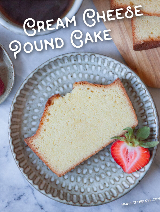 A slice of cream cheese pound cake on a plate with a strawberry garnish. There is a mug of tea next to the plate, along with a wooden serving board with the rest of the cream cheese pound cake loaf with some of it sliced up.