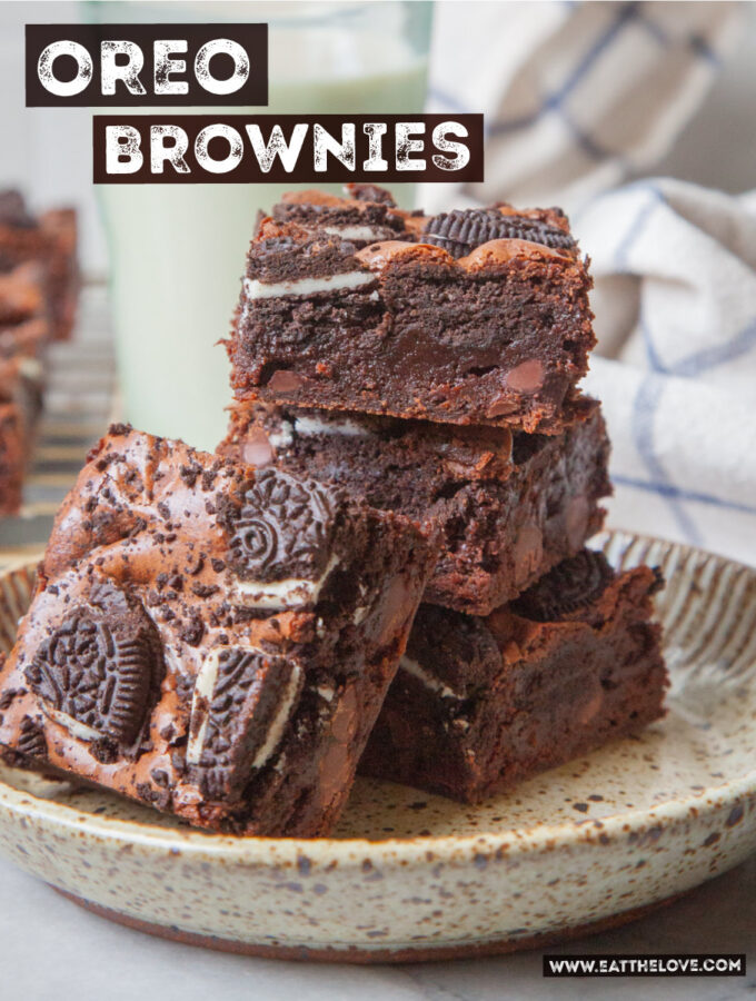A stack of Oreo brownies on a ceramic plate, with a glass of milk and more brownies on a wire cooling rack behind the stack.