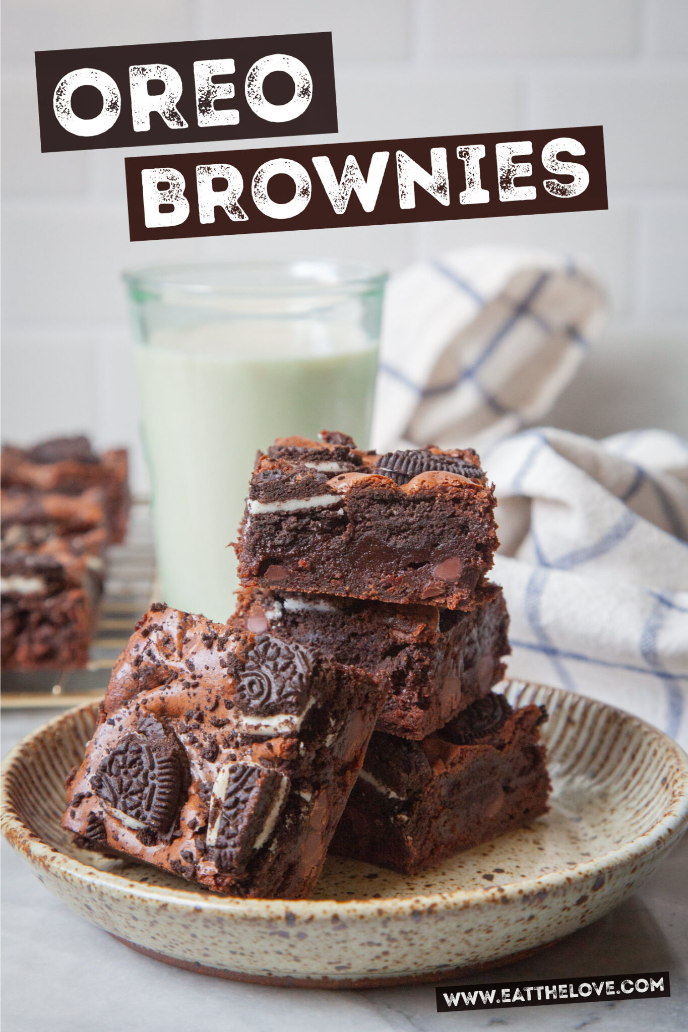 A stack of Oreo brownies on a ceramic plate, with a glass of milk and more brownies on a wire cooling rack behind the stack.