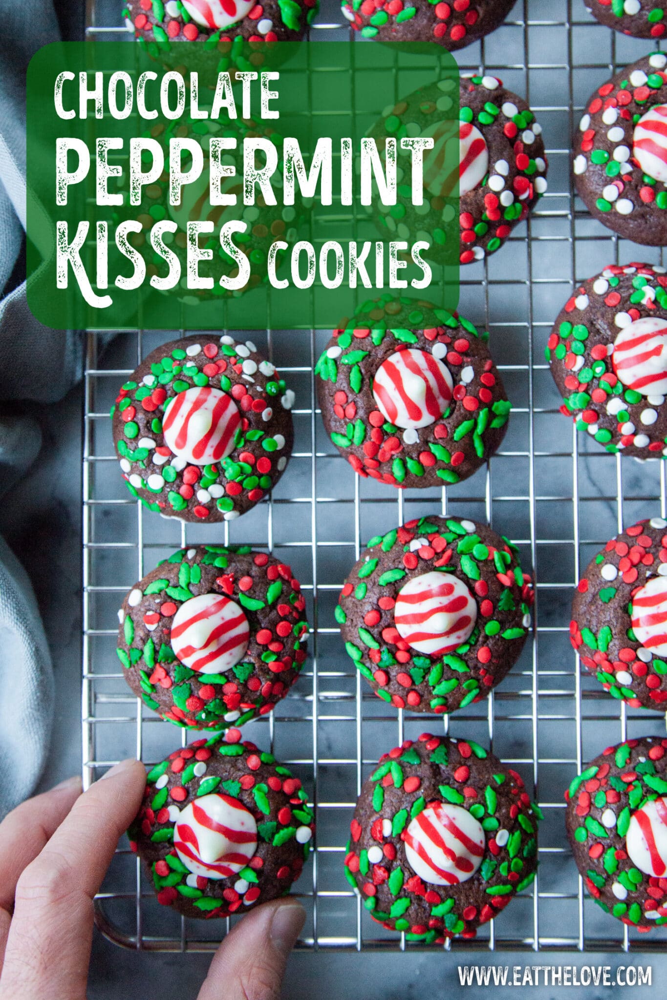 Chocolate peppermint cookies rolled in sprinkles with a peppermint kisses on top of each cookie sitting on a wire cooling rack. A hand is reaching for one of them.