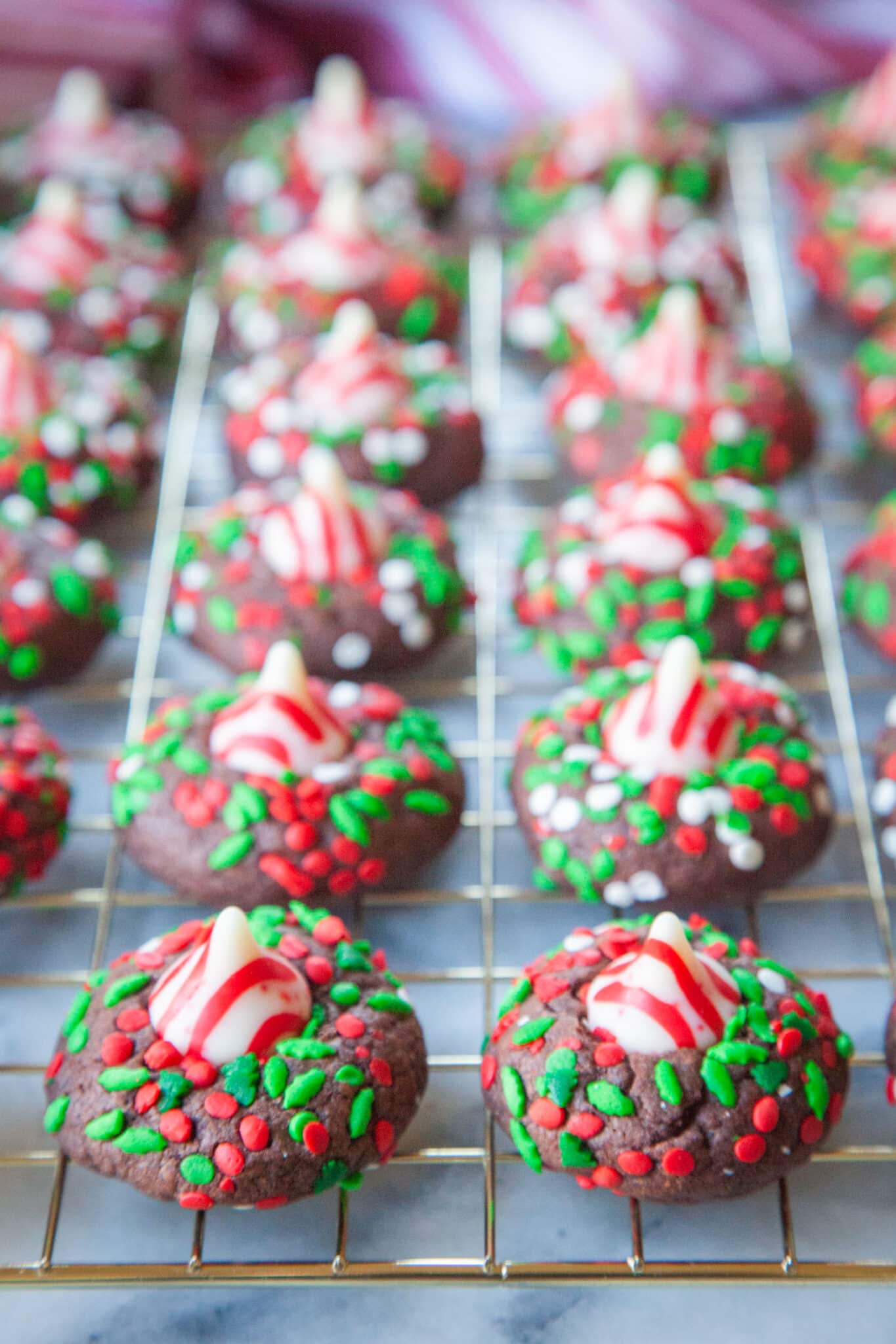 Chocolate peppermint cookies rolled in sprinkles with a peppermint kisses on top of each cookie sitting on a wire cooling rack.