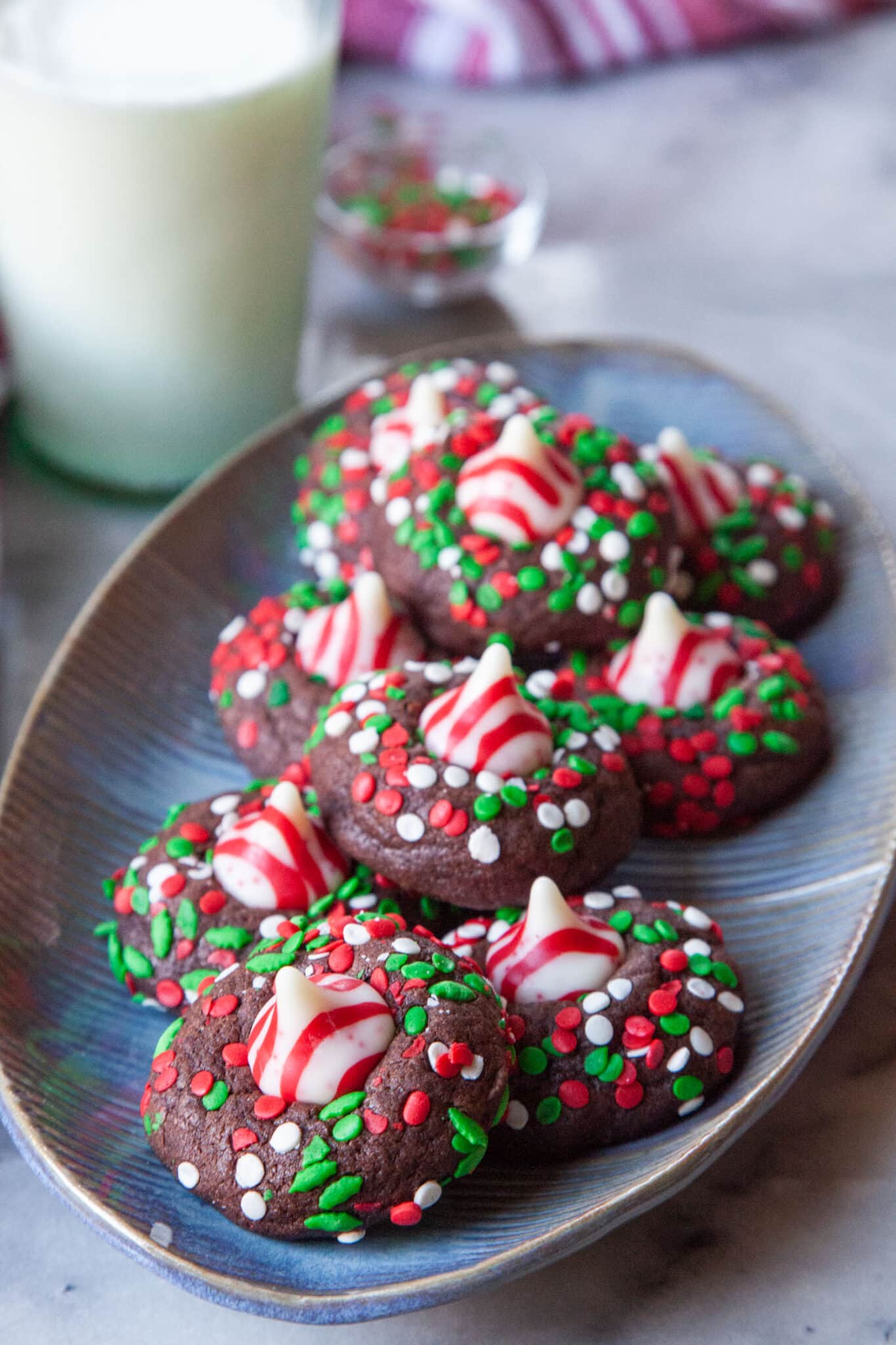 A large oval plate filled with chocolate peppermint kisses cookies. There is a glass of milk and a red and white striped cloth napkin behind the plate.
