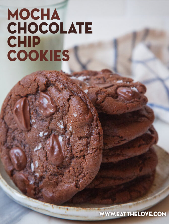 A stack of mocha chocolate chip cookies on a plate with one cookie on its side facing forward. A glass of milk and a cloth napkin is behind it.