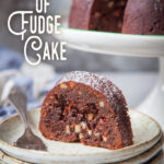 A slice of tunnel of fudge cake on a plate, with the remaining cake behind it on a cake stand.
