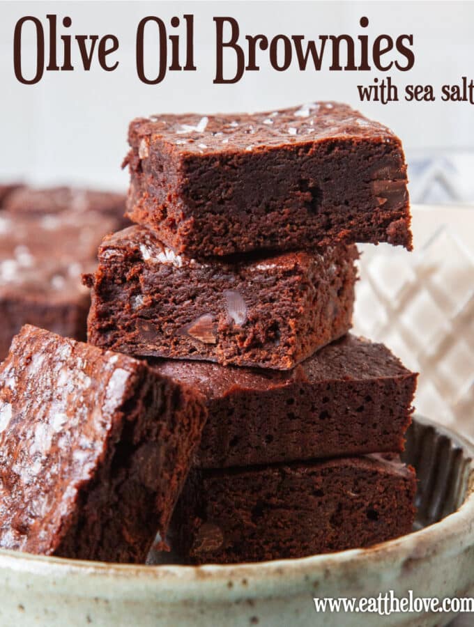 A small plate of olive oil brownies stacked on top of each other. There is a glass of milk and more brownies behind the plate.