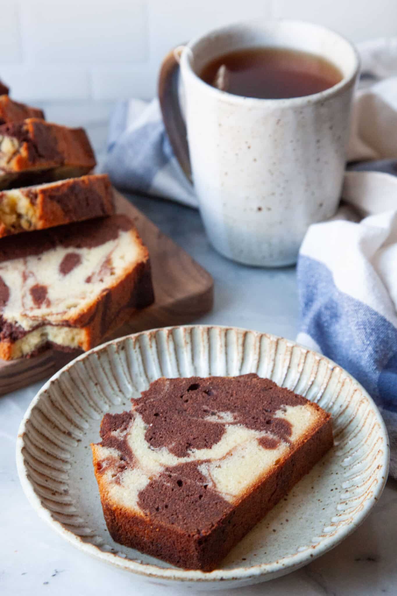 A slice of marbled pound cake on a plate. Behind the cake slice is a mug of tea and the rest of the cake on a cutting board.