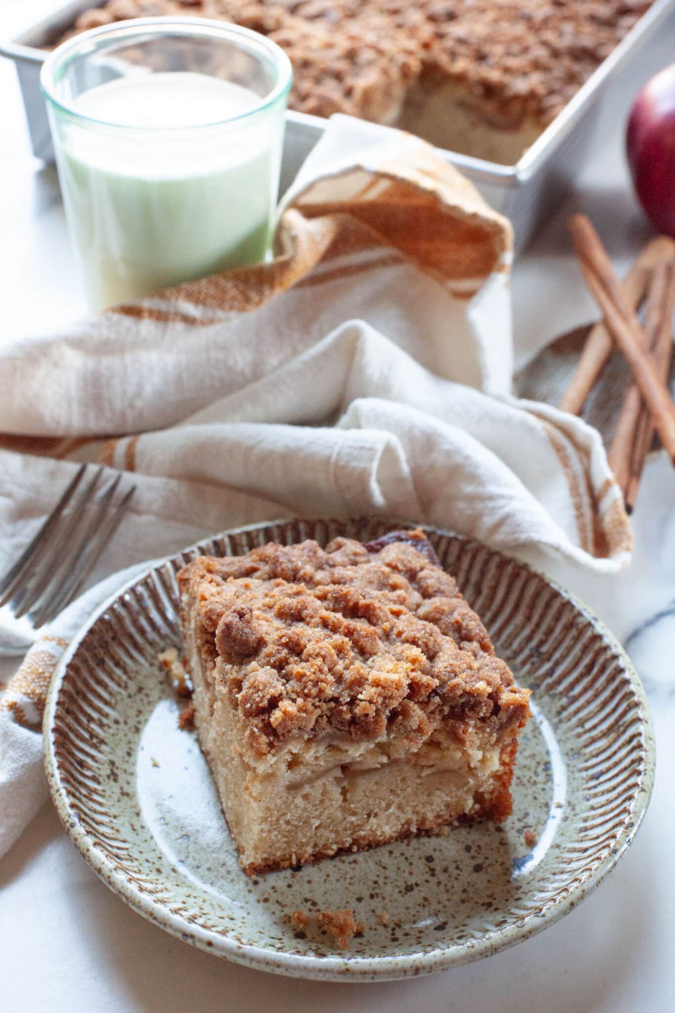 A slice of apple coffee cake on a plate, with a glass of milk and the remaining cake behind the plate.