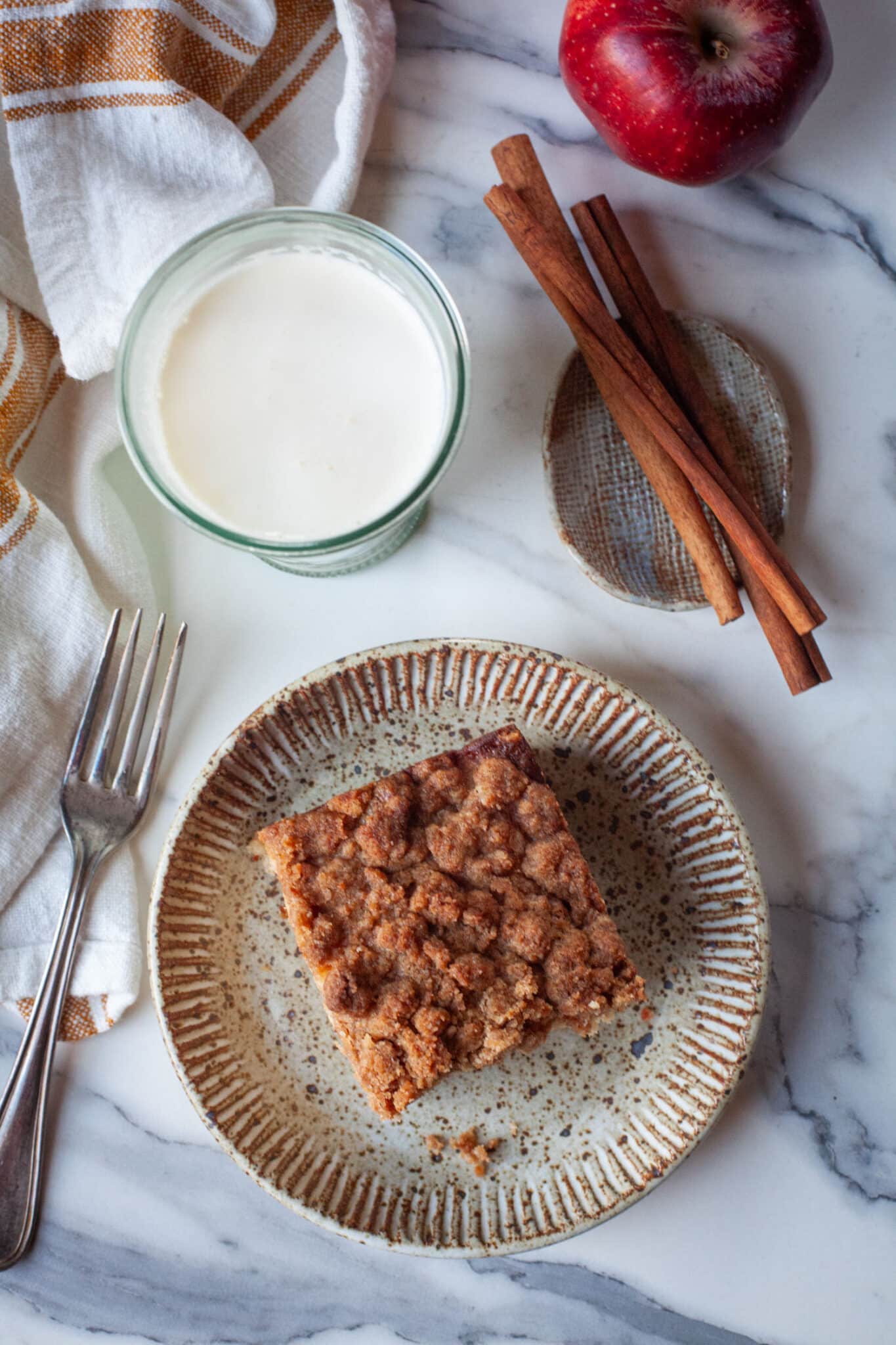 A slice of apple coffee cake with a glass of milk, cinnamon sticks and an apple next to it.