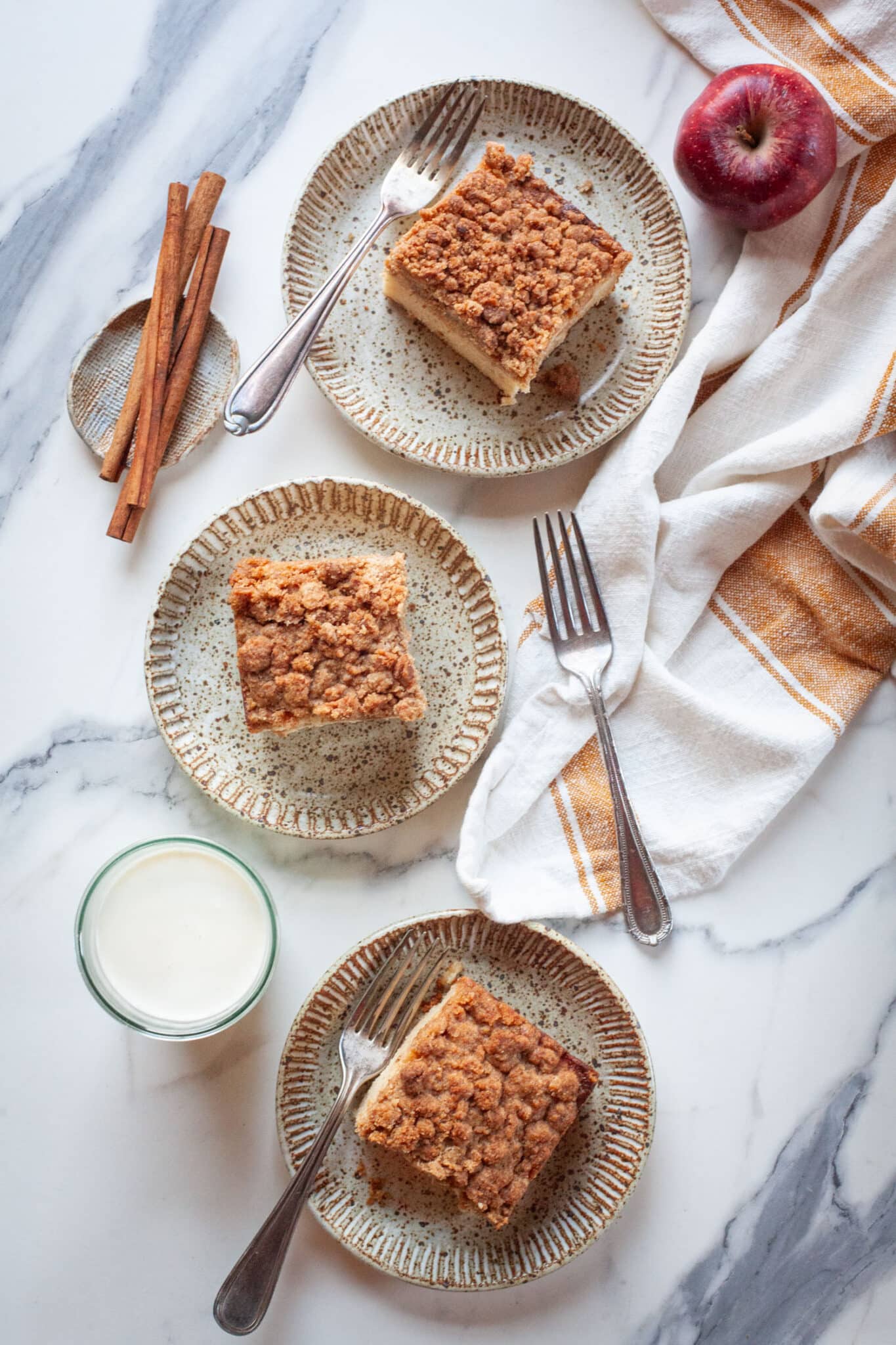 Three slices of apple coffee cake on three different plates, with a glass of milk, cinnamon sticks and an apple next to the plates.