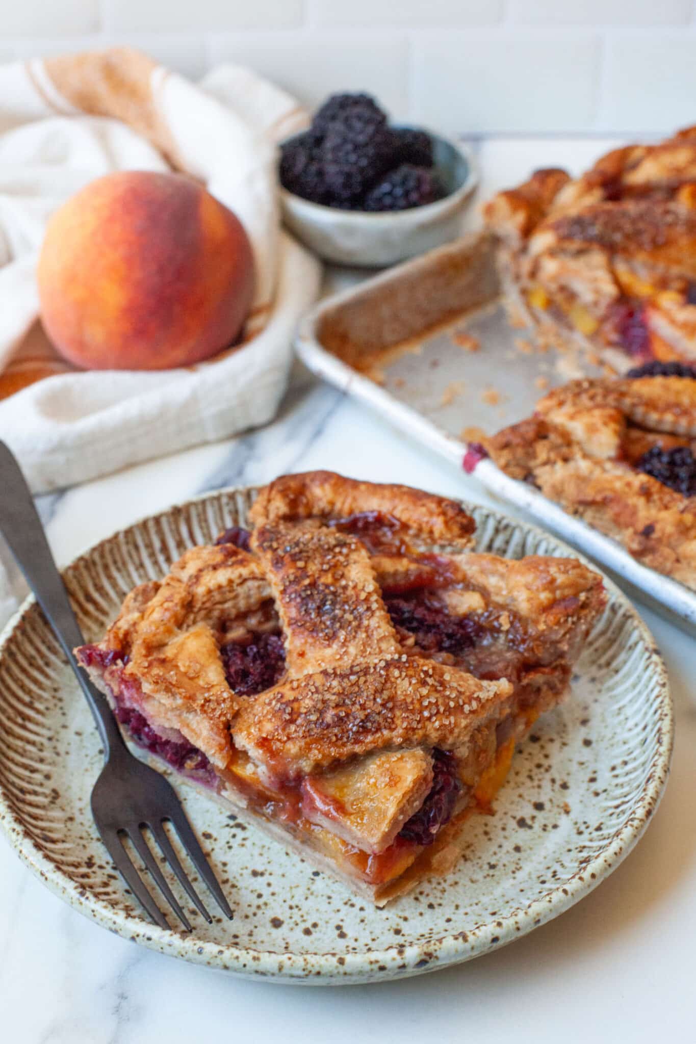 A slice of peach and blackberry slab pie on a plate, with a peach and blackberries behind it, along with the remaining pie.