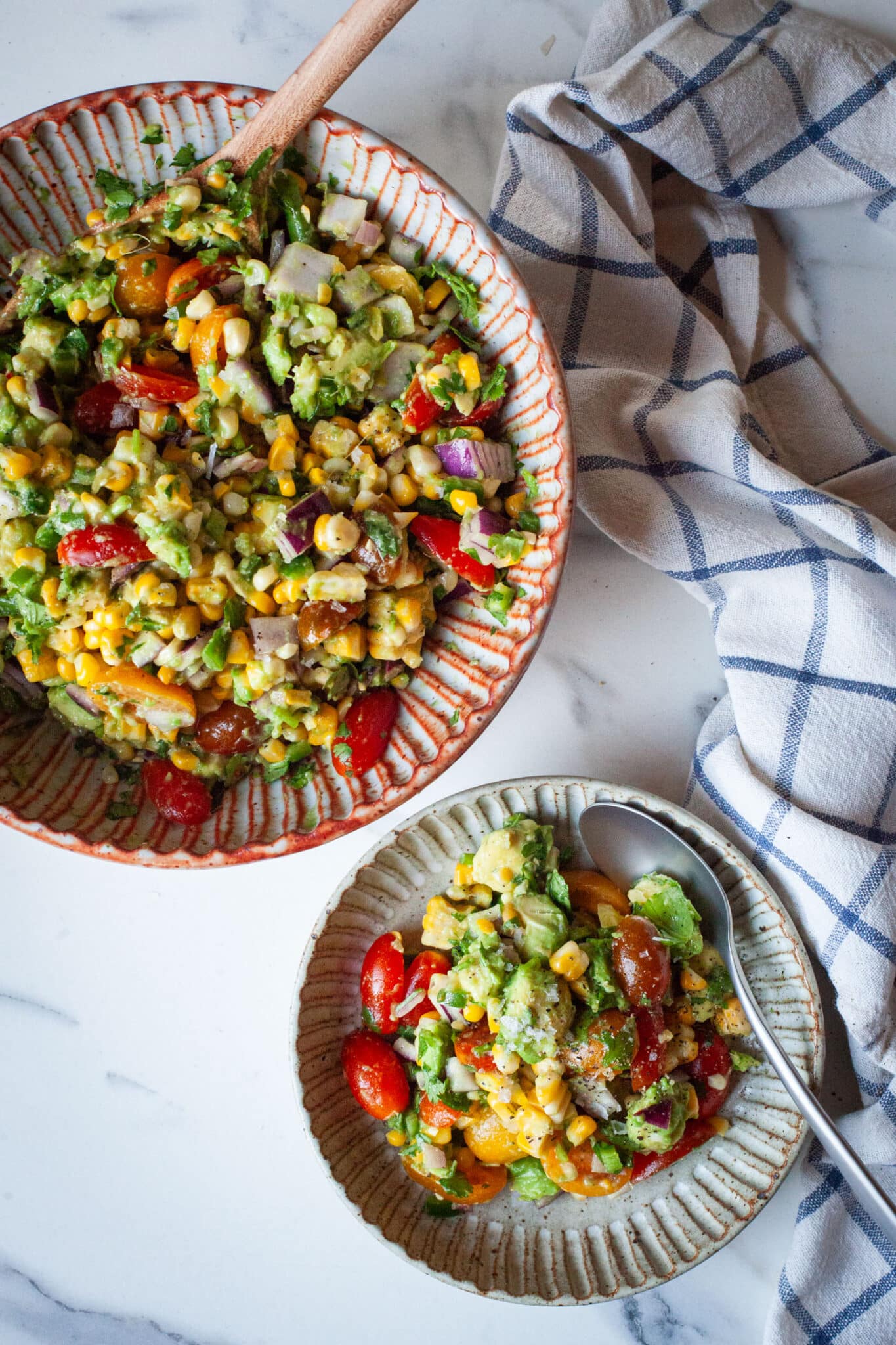 A bowl of avocado and corn salad with tomatoes and lime dressing. There is a larger bowl of the salad next to it.
