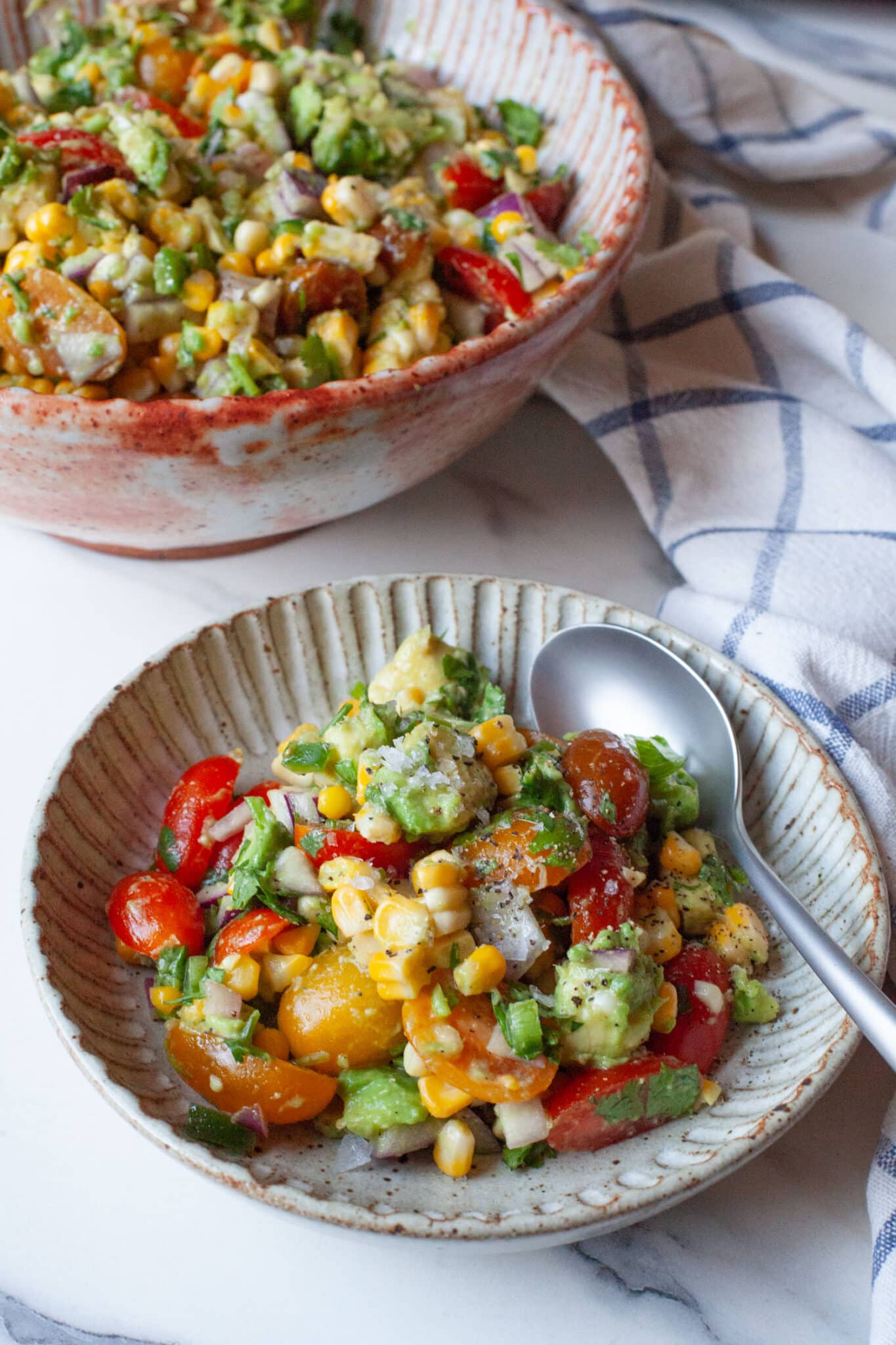 A bowl of avocado and corn salad with tomatoes and lime dressing. There is a larger bowl of the salad behind it.