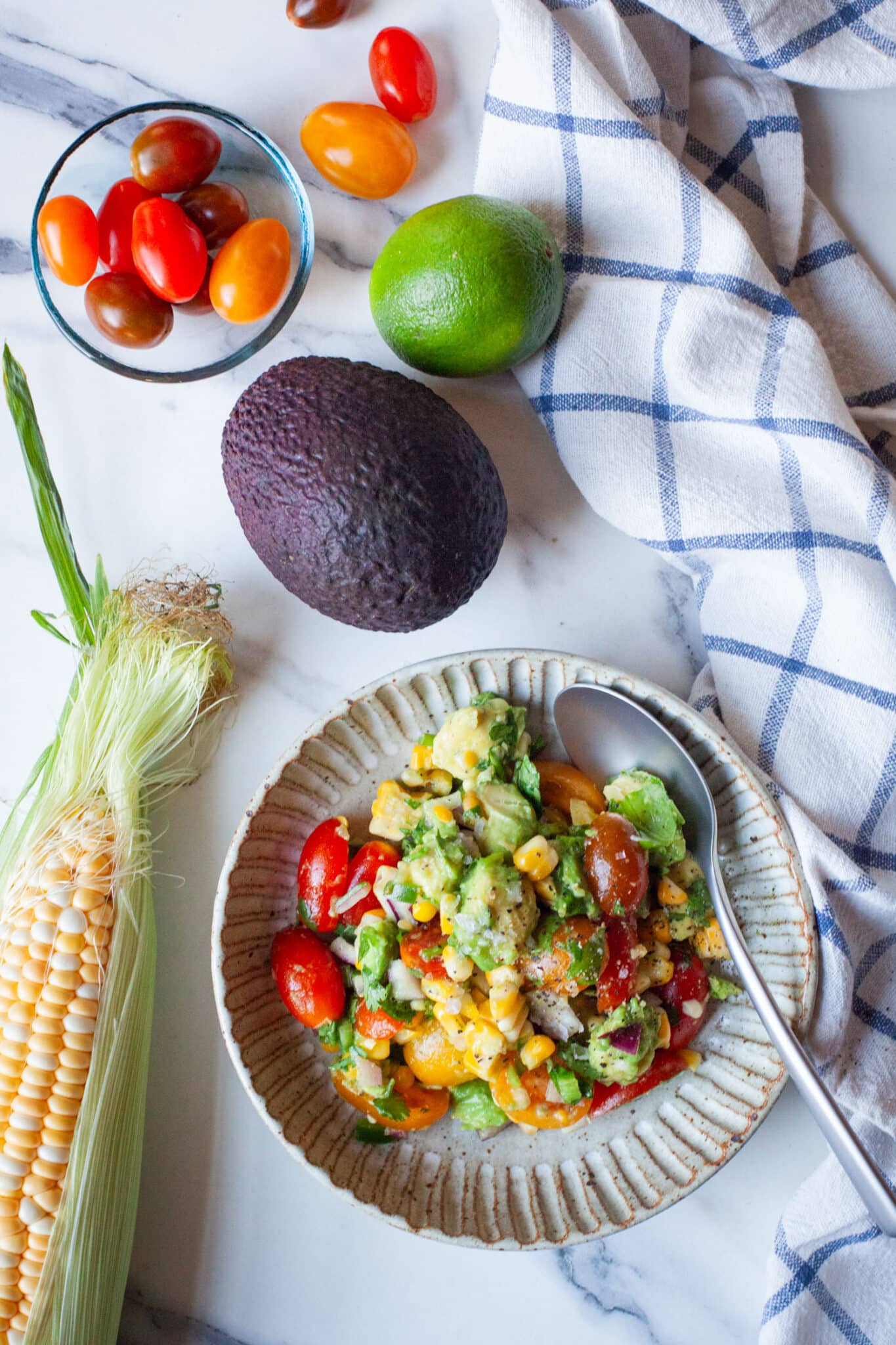 A bowl of avocado and corn salad, with an ear of corn, grape tomatoes, and a lime next to the bowl.