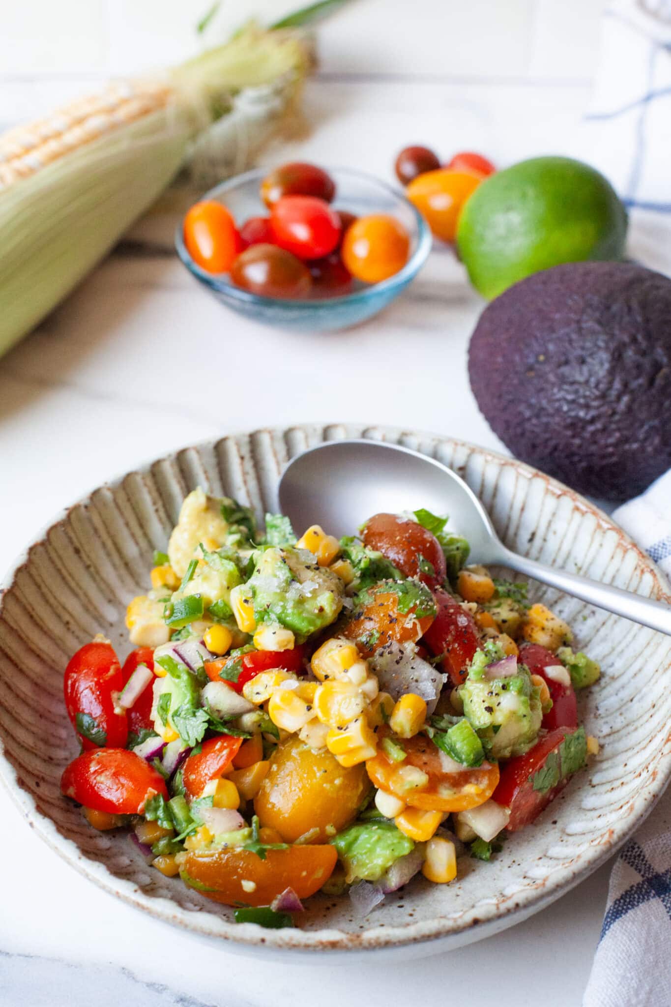 A bowl of avocado and corn salad, with an ear of corn, grape tomatoes, and a lime behind the bowl.