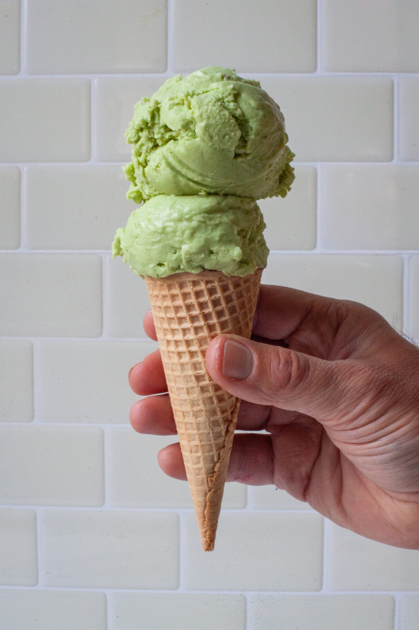 A hand holding a cone with two scoops of avocado and lime ice cream in it.
