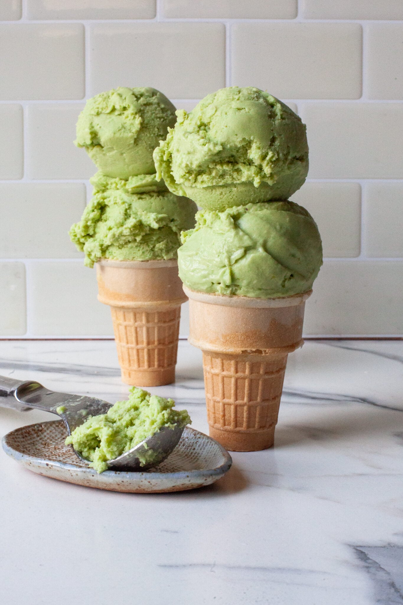 Two ice cream cones with scoops of avocado and lime ice cream in them sitting on a marble table, with an ice cream scoop with more ice cream in it next to the cones.