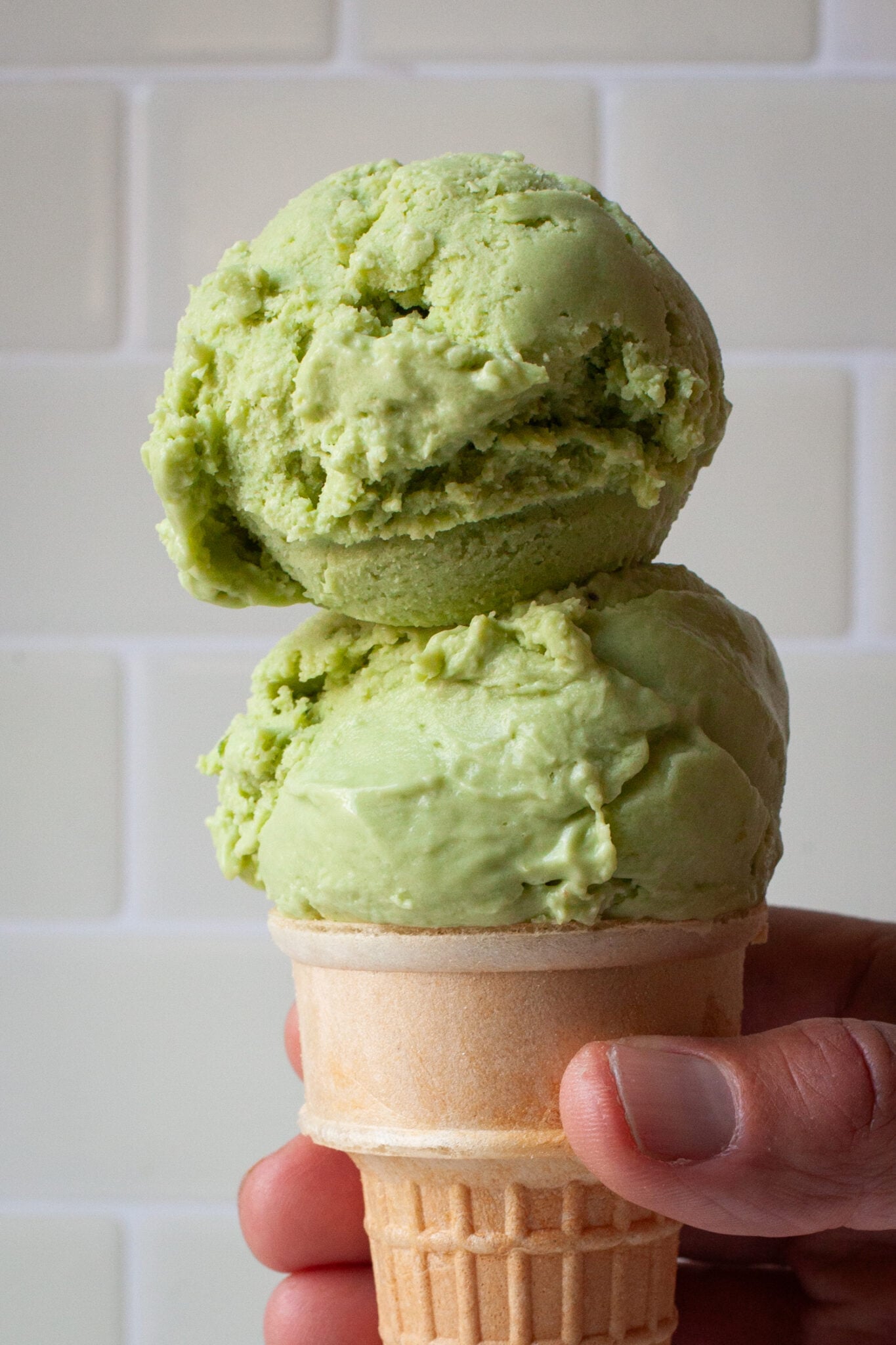 A close up of two scoops of avocado and lime ice cream in a cake cone being held by a hand.