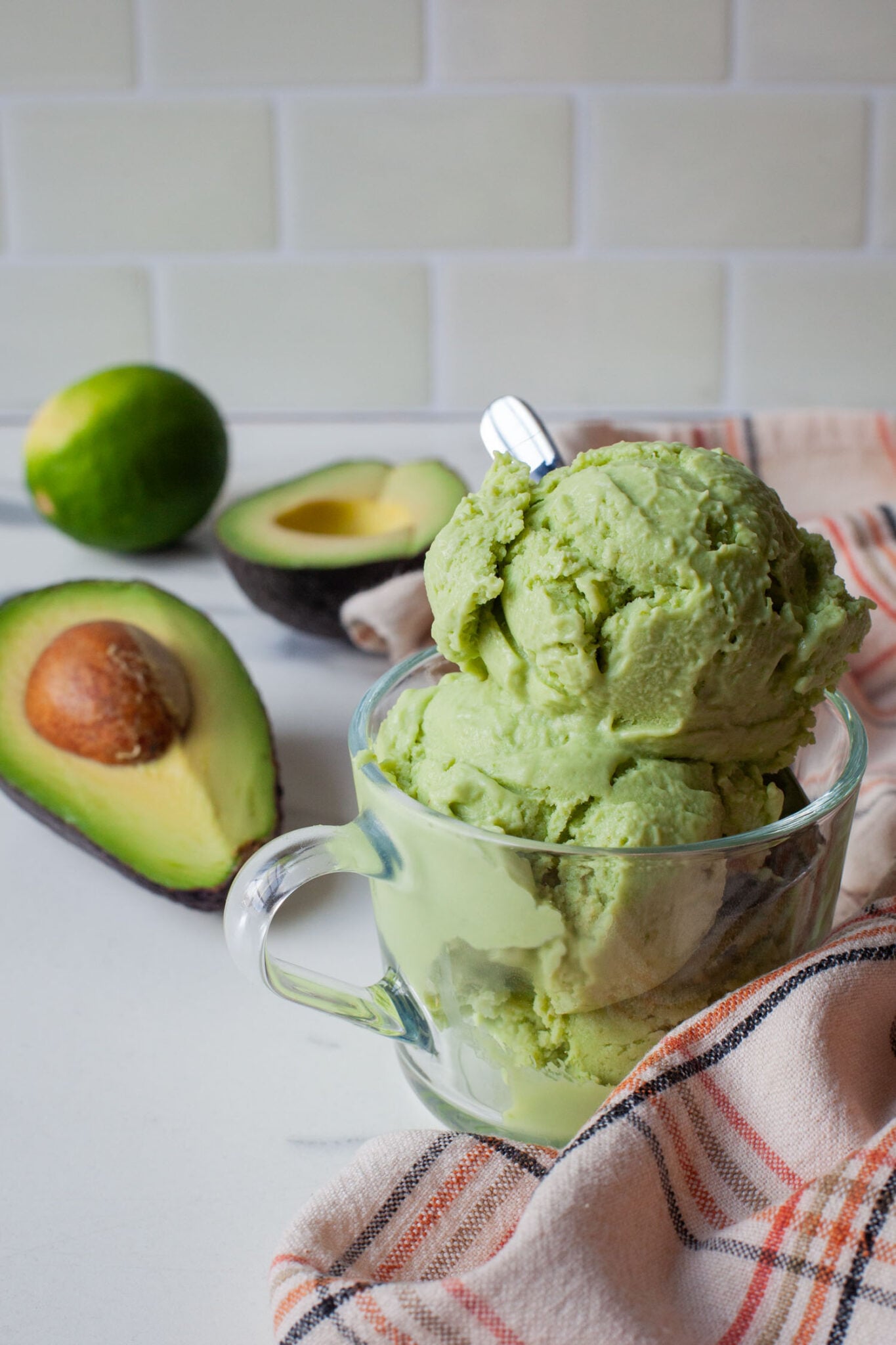 A scoop of avocado and lime ice cream in a glass cup, with a cut avocado and lime behind it.