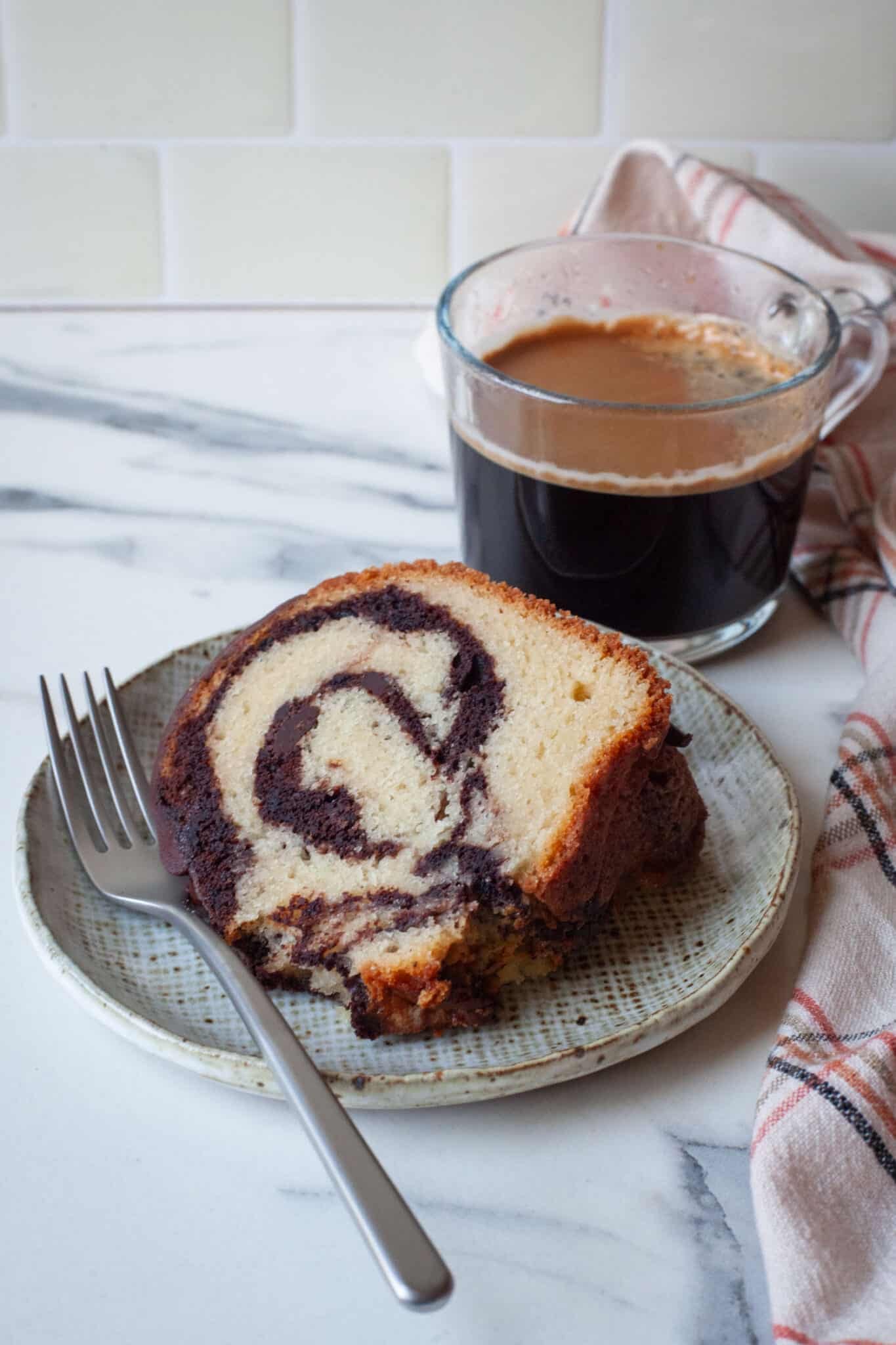 A slice of marble burnt cake on a plate, with an Americano coffee behind it.